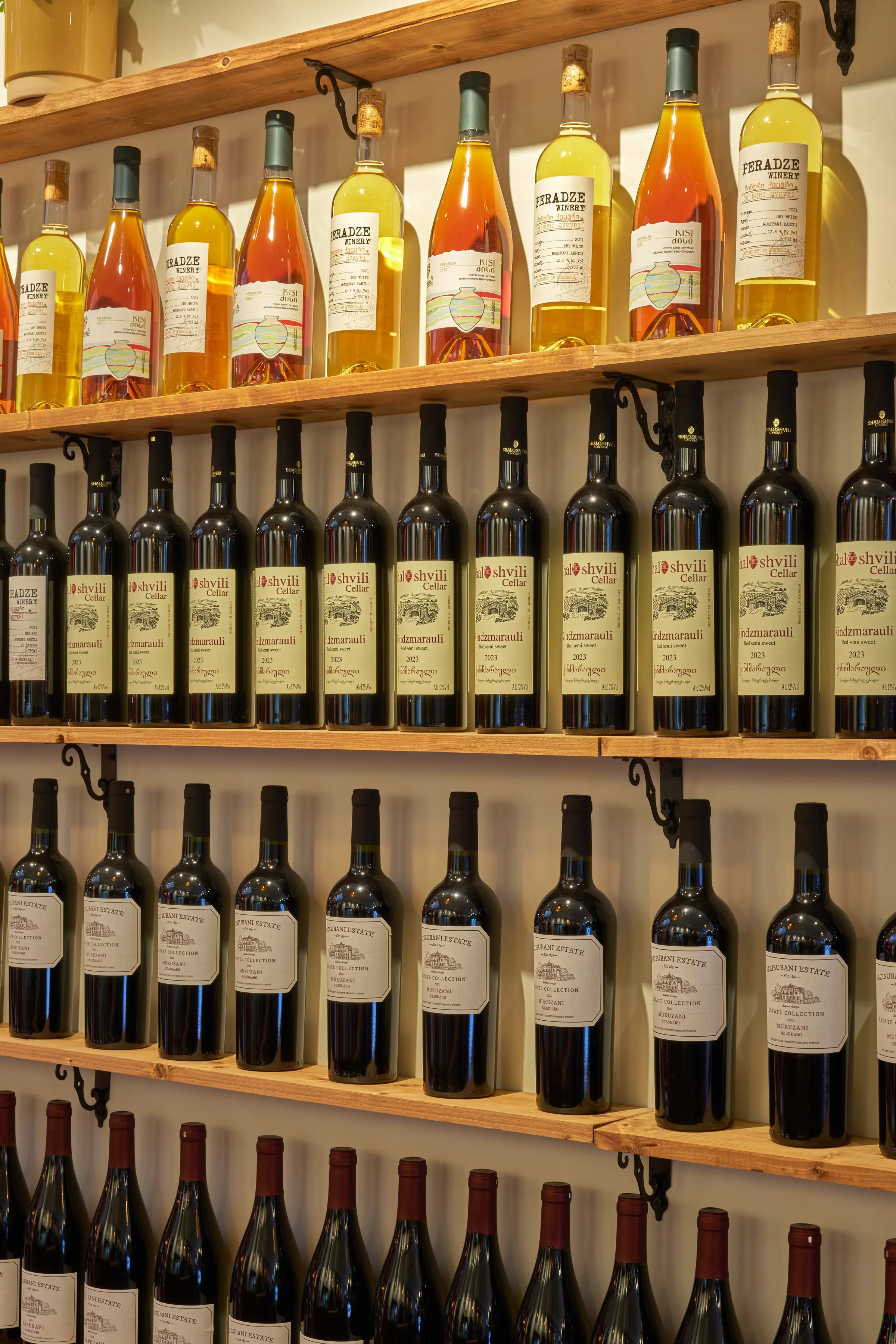 Detalle de botellas de vino ámbar y tinto en estantes de madera, con etiquetas de Peradze, Shaloshvili y Vazisubani